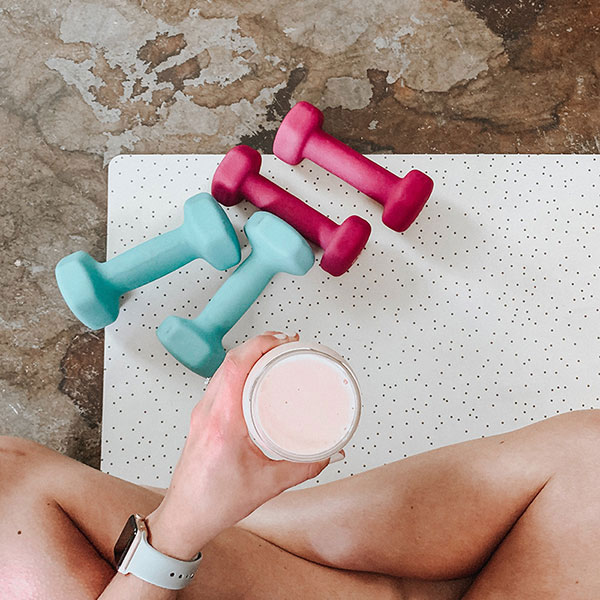 Žena sedící na podložce s činkami, držící smoothie.